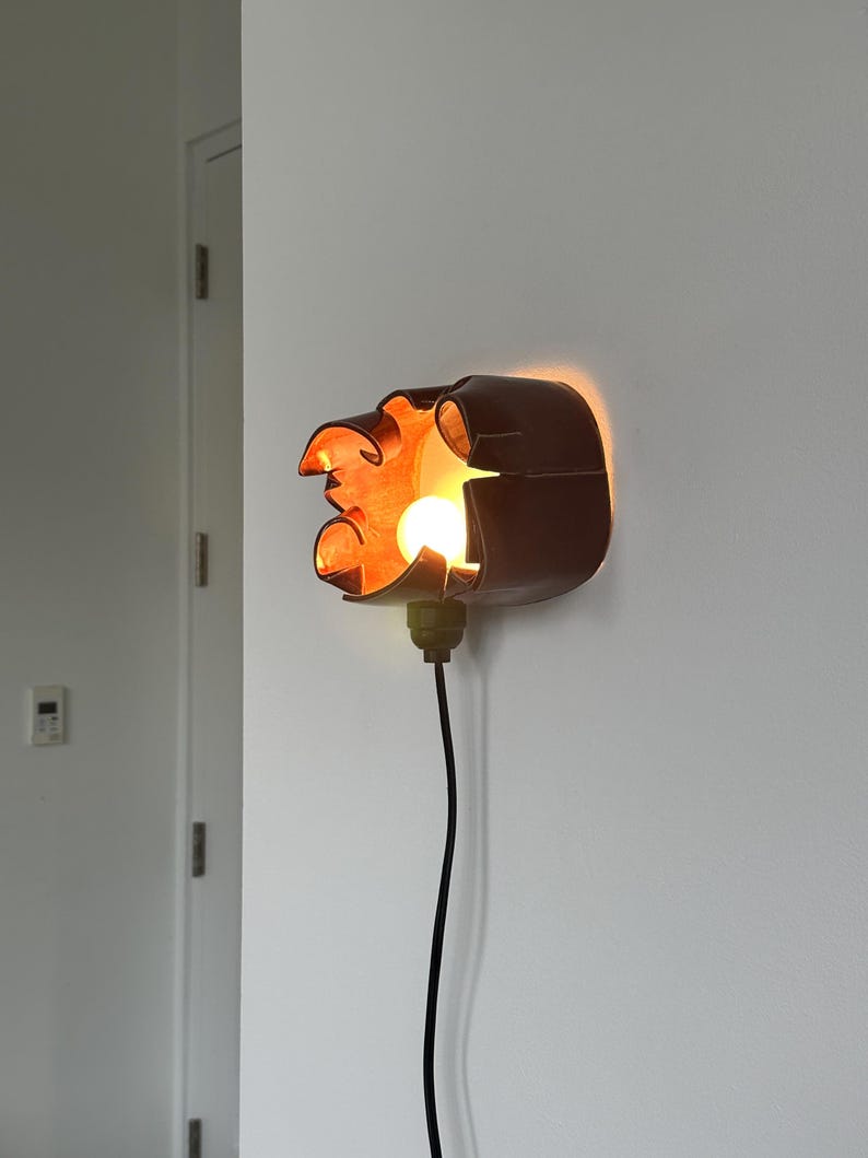 May include: A wall-mounted, brown, decorative light fixture with an orange glow. The fixture has an abstract, organic shape and a visible light bulb. A black cord extends downward from the fixture.