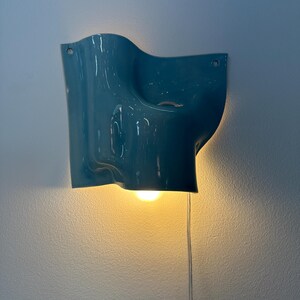 May include: A glossy teal wall sconce with an organic, undulating shape. The fixture features a central opening that emits a warm, downward-facing light from an internal bulb. A thin, transparent power cord hangs vertically from the bottom edge.