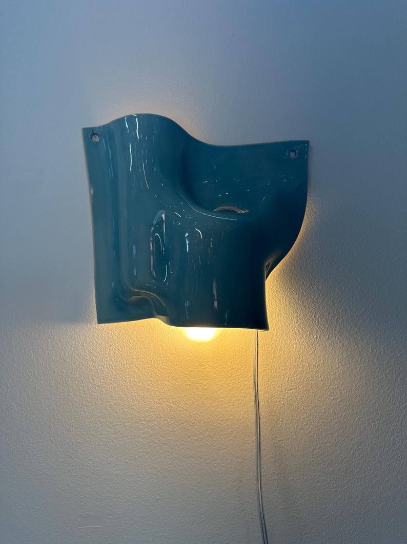 May include: A glossy teal wall sconce with an organic, undulating shape. The fixture features a central opening that emits a warm, downward-facing light from an internal bulb. A thin, transparent power cord hangs vertically from the bottom edge.