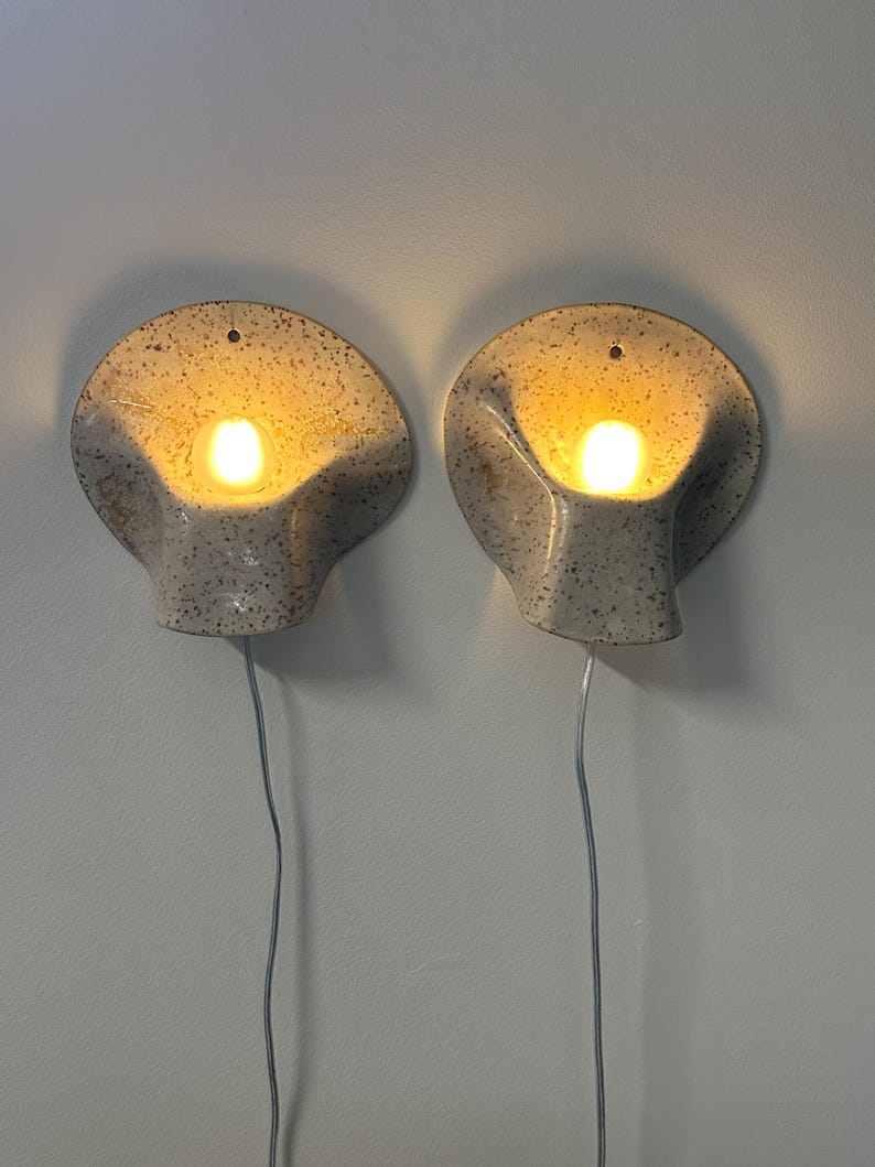 May include: A pair of wall-mounted sconces featuring a speckled off-white ceramic shell design. Each lamp emits a warm, glowing light from a central bulb and includes a thin, clear electrical cord extending downwards from the base of the fixture.