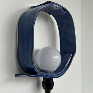 Puede incluir: Aplique de pared de cer&aacute;mica azul marino con acabado brillante. La l&aacute;mpara tiene un dise&ntilde;o curvo &uacute;nico que rodea una bombilla blanca. La l&aacute;mpara est&aacute; montada en una pared blanca y tiene un cable negro.