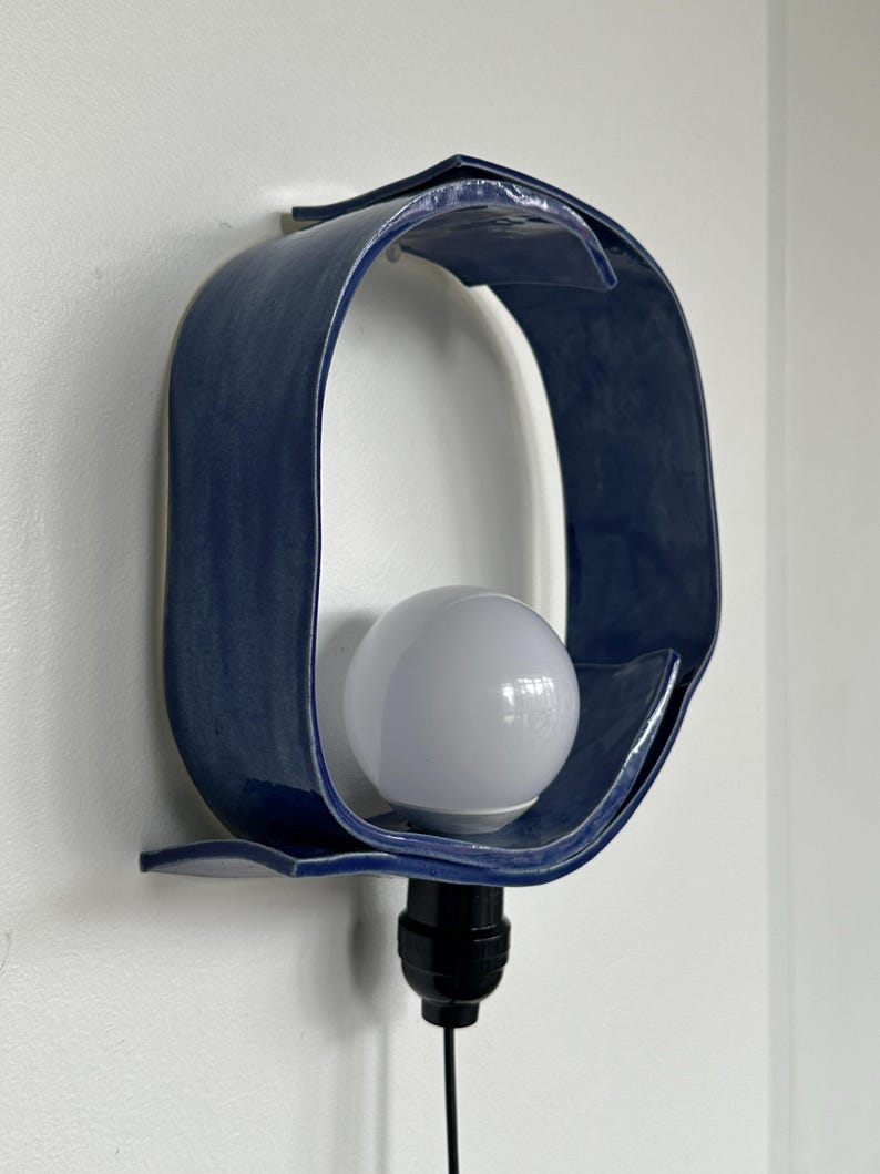 Puede incluir: Aplique de pared de cer&aacute;mica azul marino con acabado brillante. La l&aacute;mpara tiene un dise&ntilde;o curvo &uacute;nico que rodea una bombilla blanca. La l&aacute;mpara est&aacute; montada en una pared blanca y tiene un cable negro.