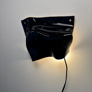 Puede incluir: Una l&aacute;mpara de pared con una pantalla de cer&aacute;mica negra brillante y ondulada que recuerda a una tela plegada. El aplique cuenta con dos peque&ntilde;os orificios circulares de montaje en las esquinas superiores y emite luz c&aacute;lida.