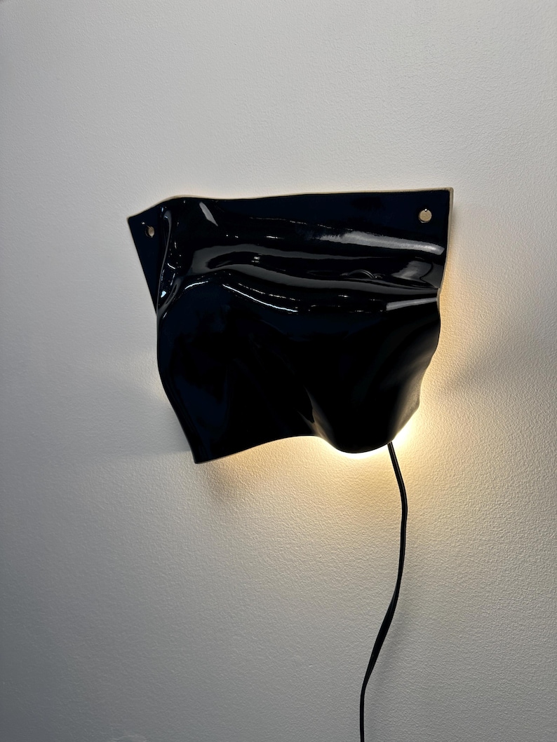Puede incluir: Una l&aacute;mpara de pared con una pantalla de cer&aacute;mica negra brillante y ondulada que recuerda a una tela plegada. El aplique cuenta con dos peque&ntilde;os orificios circulares de montaje en las esquinas superiores y emite luz c&aacute;lida.