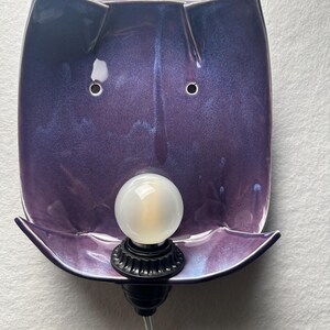 May include: A wall-mounted lamp featuring a glossy, deep purple ceramic shade with a curved, rectangular shape. The shade has two small circular holes near the top and a black socket at the bottom holding a round, frosted white light bulb.