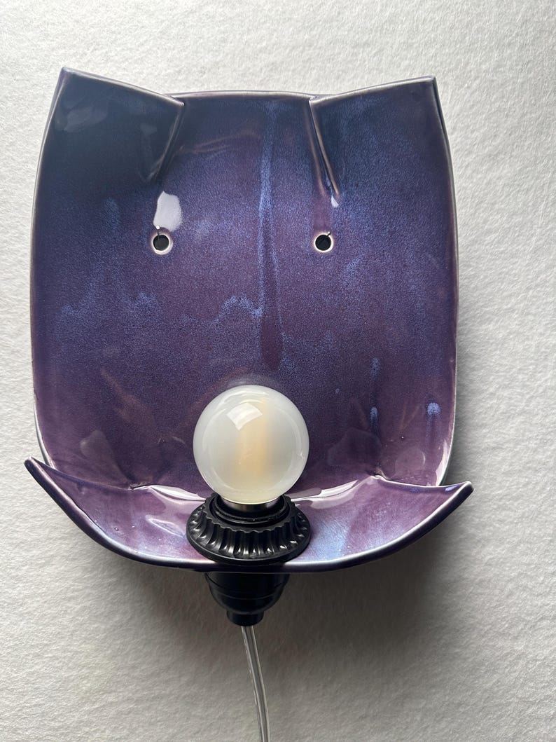 May include: A wall-mounted lamp featuring a glossy, deep purple ceramic shade with a curved, rectangular shape. The shade has two small circular holes near the top and a black socket at the bottom holding a round, frosted white light bulb.