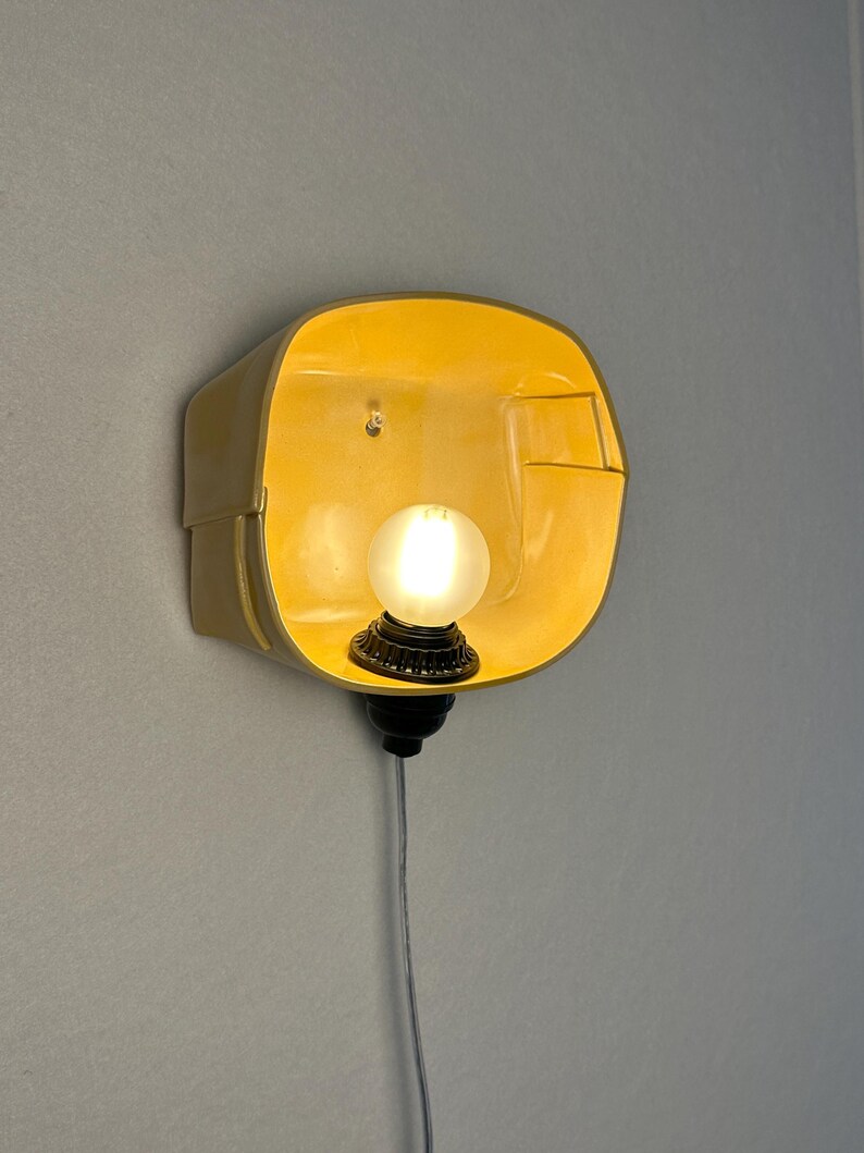 May include: A wall-mounted lamp featuring a bright yellow, rounded rectangular shade with a smooth finish. A small, frosted spherical light bulb is centered within the shade, attached to a black socket base with a thin, light-colored power cord.