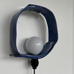 Puede incluir: Una l&aacute;mpara de pared de cer&aacute;mica azul marino con un dise&ntilde;o curvo y abierto. La l&aacute;mpara contiene una bombilla blanca y tiene un cable negro. La l&aacute;mpara est&aacute; montada en una pared blanca.