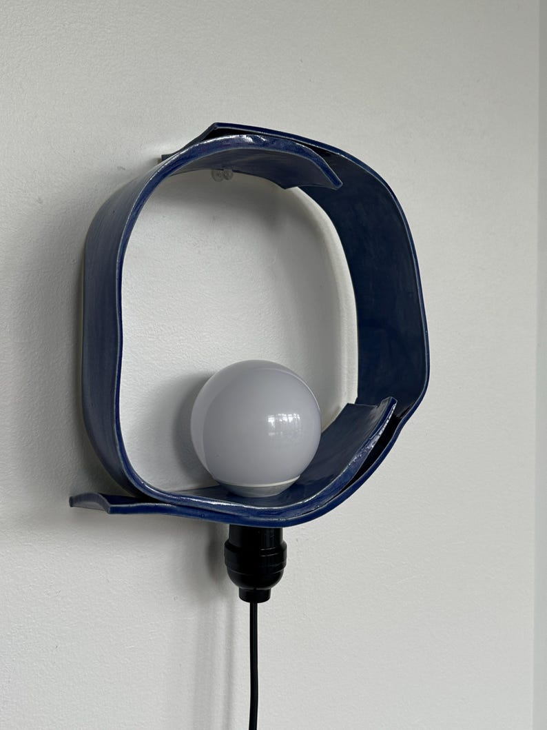 Puede incluir: Una l&aacute;mpara de pared de cer&aacute;mica azul marino con un dise&ntilde;o curvo y abierto. La l&aacute;mpara contiene una bombilla blanca y tiene un cable negro. La l&aacute;mpara est&aacute; montada en una pared blanca.