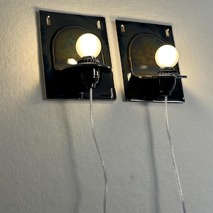May include: Two wall sconces with a dark blue and gold finish. Each sconce features a black base with a round, glowing light bulb. The sconces are mounted on a light-colored wall, with the electrical cords visible.