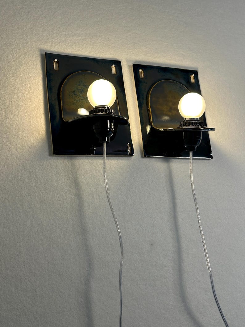 May include: Two wall sconces with a dark blue and gold finish. Each sconce features a black base with a round, glowing light bulb. The sconces are mounted on a light-colored wall, with the electrical cords visible.