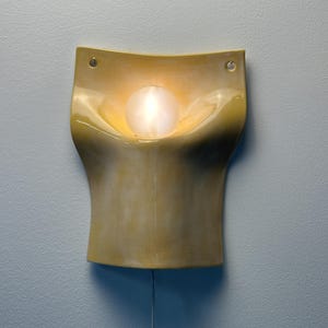 May include: A wall-mounted sconce featuring a curved, glossy yellow ceramic body with a smooth, organic shape. A single round, frosted glass bulb is centered in the recessed upper section, emitting a warm glow. Two small holes are visible at the top corners.