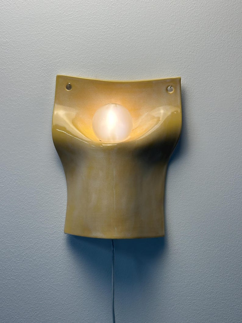 May include: A wall-mounted sconce featuring a curved, glossy yellow ceramic body with a smooth, organic shape. A single round, frosted glass bulb is centered in the recessed upper section, emitting a warm glow. Two small holes are visible at the top corners.