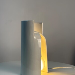May include: A white ceramic table lamp with a smooth, curved, cylindrical structure that features a vertical cutout. The lamp emits a warm, soft glow from an internal light bulb visible through the opening, and includes a thin white power cord.