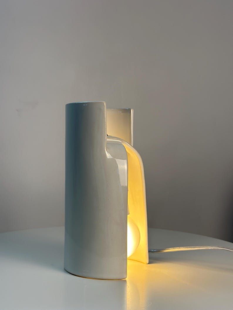 May include: A white ceramic table lamp with a smooth, curved, cylindrical structure that features a vertical cutout. The lamp emits a warm, soft glow from an internal light bulb visible through the opening, and includes a thin white power cord.