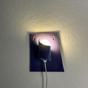 Pode incluir: Uma arandela de parede moderna com um design geom&eacute;trico azul escuro. A arandela apresenta uma sombra cil&iacute;ndrica e uma l&acirc;mpada redonda e brilhante. A lumin&aacute;ria &eacute; montada em uma placa retangular com um efeito gradiente, e um cabo prateado se estende para baixo.