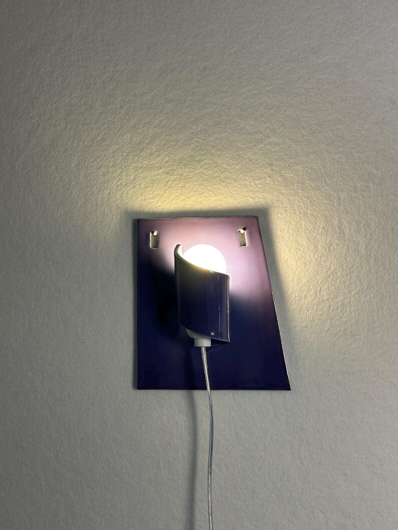 Pode incluir: Uma arandela de parede moderna com um design geom&eacute;trico azul escuro. A arandela apresenta uma sombra cil&iacute;ndrica e uma l&acirc;mpada redonda e brilhante. A lumin&aacute;ria &eacute; montada em uma placa retangular com um efeito gradiente, e um cabo prateado se estende para baixo.