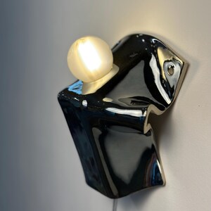 May include: A wall-mounted light fixture featuring a glossy black ceramic base with an irregular, folded shape. A single round, frosted glass light bulb is seated in a circular opening at the top, emitting a warm, soft glow against the wall.