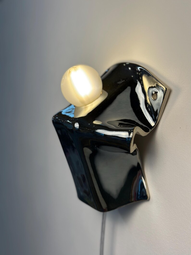 May include: A wall-mounted light fixture featuring a glossy black ceramic base with an irregular, folded shape. A single round, frosted glass light bulb is seated in a circular opening at the top, emitting a warm, soft glow against the wall.