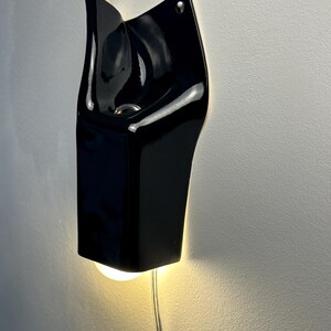 May include: A glossy black ceramic wall sconce with an organic, folded silhouette. The fixture features a central opening housing a light bulb that emits a warm glow downward, with a thin, transparent power cord extending from the bottom edge.