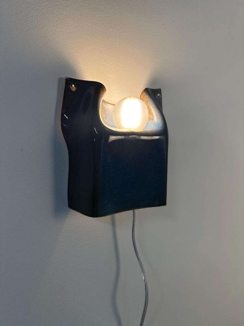 May include: A dark blue ceramic wall sconce with a glossy finish and a curved, organic shape. The fixture features a central cutout holding a single glowing white spherical light bulb, with a thin power cord extending downward from the base.