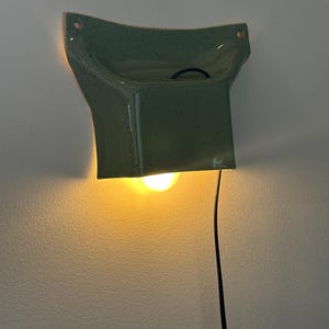 May include: A wall-mounted ceramic lamp with a speckled sage green glaze. The fixture has a flared, angular shape with two mounting holes at the top corners. A warm light glows from beneath the structure, and a thin black power cord hangs down.