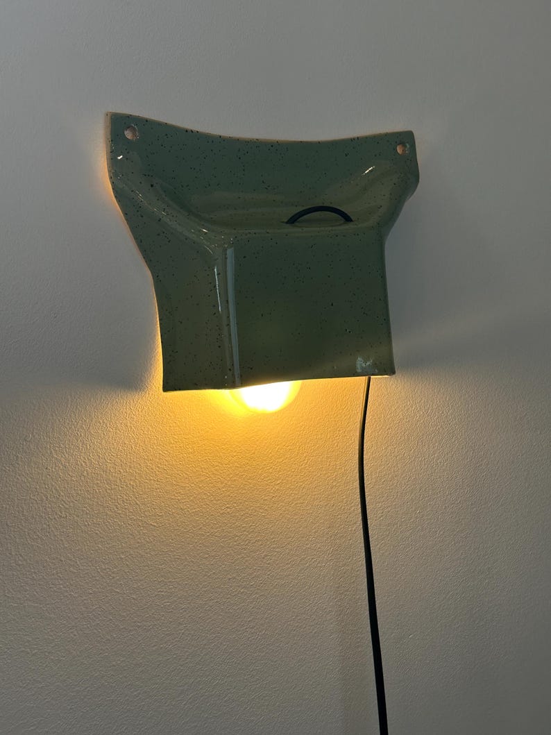 May include: A wall-mounted ceramic lamp with a speckled sage green glaze. The fixture has a flared, angular shape with two mounting holes at the top corners. A warm light glows from beneath the structure, and a thin black power cord hangs down.