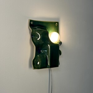May include: A wall-mounted ceramic light fixture with a glossy dark green finish and an irregular, undulating surface. The piece features a circular cutout holding a glowing white spherical bulb and includes two small holes for mounting hardware.