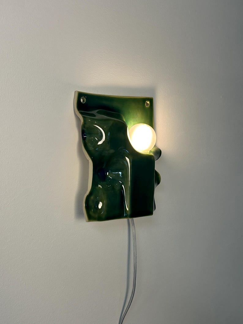 May include: A wall-mounted ceramic light fixture with a glossy dark green finish and an irregular, undulating surface. The piece features a circular cutout holding a glowing white spherical bulb and includes two small holes for mounting hardware.