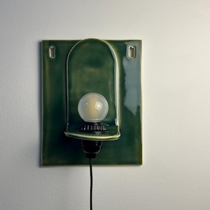 May include: A wall-mounted sconce featuring a rectangular, glossy forest green ceramic backplate with a central arched indentation. A black socket holds a round, frosted white light bulb, and a black power cord extends from the bottom.
