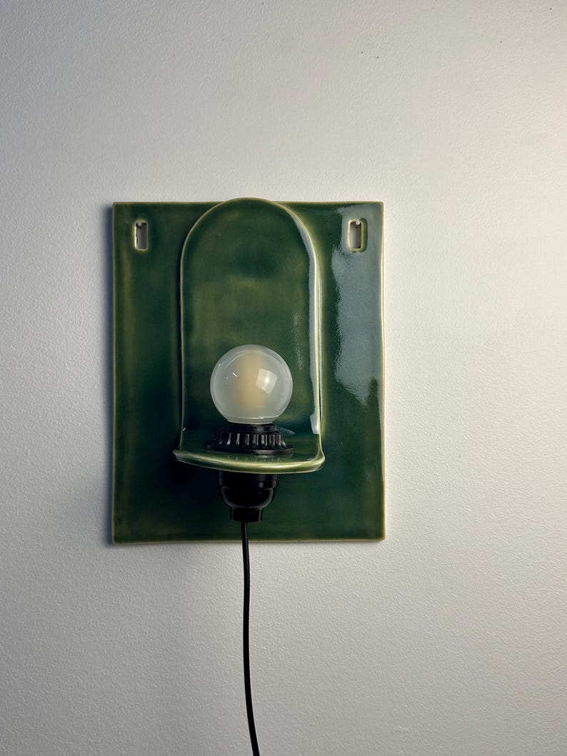 May include: A wall-mounted sconce featuring a rectangular, glossy forest green ceramic backplate with a central arched indentation. A black socket holds a round, frosted white light bulb, and a black power cord extends from the bottom.