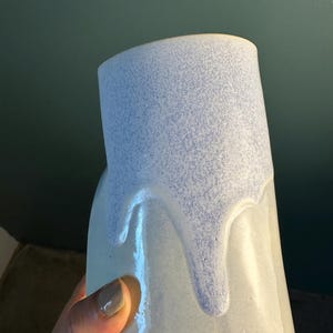 K&ouml;nnte beinhalten: Eine Keramikvase mit einer glatten, cremewei&szlig;en Grundglasur und einer strukturierten, gesprenkelten hellblauen Glasur, die vom oberen Rand herabl&auml;uft. Das Gef&auml;&szlig; hat eine zylindrische Form mit einer leicht unregelm&auml;&szlig;igen, organischen Silhouette.
