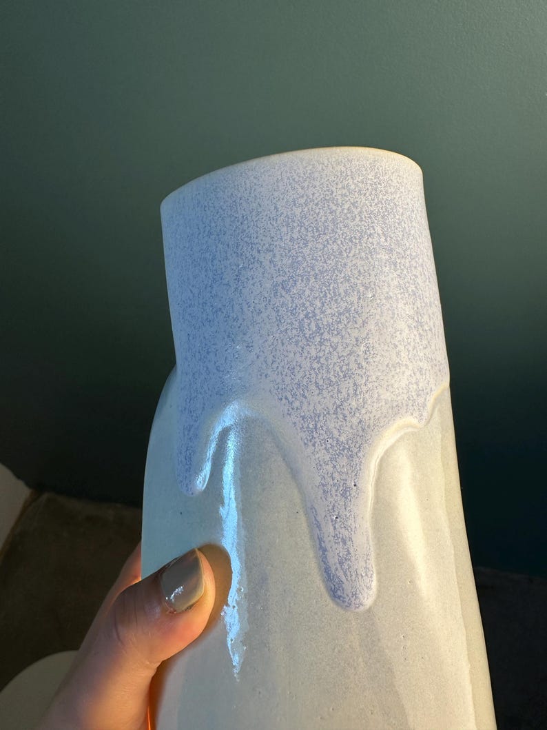 K&ouml;nnte beinhalten: Eine Keramikvase mit einer glatten, cremewei&szlig;en Grundglasur und einer strukturierten, gesprenkelten hellblauen Glasur, die vom oberen Rand herabl&auml;uft. Das Gef&auml;&szlig; hat eine zylindrische Form mit einer leicht unregelm&auml;&szlig;igen, organischen Silhouette.