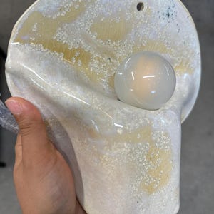 May include: A wall-mounted ceramic lamp featuring a curved, organic shape with a glossy white and pale yellow crackle glaze finish. The fixture includes a single circular opening for mounting and a central, spherical white glass light bulb.
