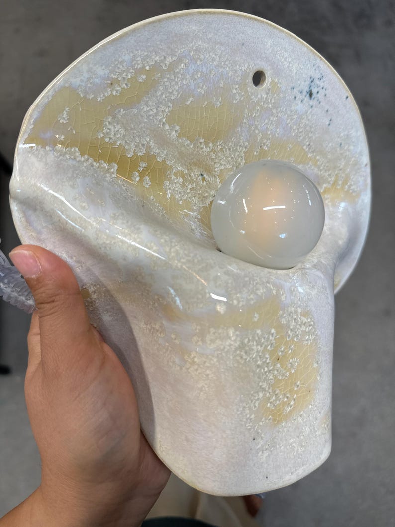 May include: A wall-mounted ceramic lamp featuring a curved, organic shape with a glossy white and pale yellow crackle glaze finish. The fixture includes a single circular opening for mounting and a central, spherical white glass light bulb.