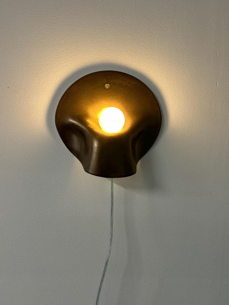May include: A wall-mounted lamp featuring a dark, metallic, circular shade with a fluid, organic, shell-like shape. A single, bright, warm-toned light bulb is centered within the recessed portion of the shade, with a thin, clear power cord extending downward.