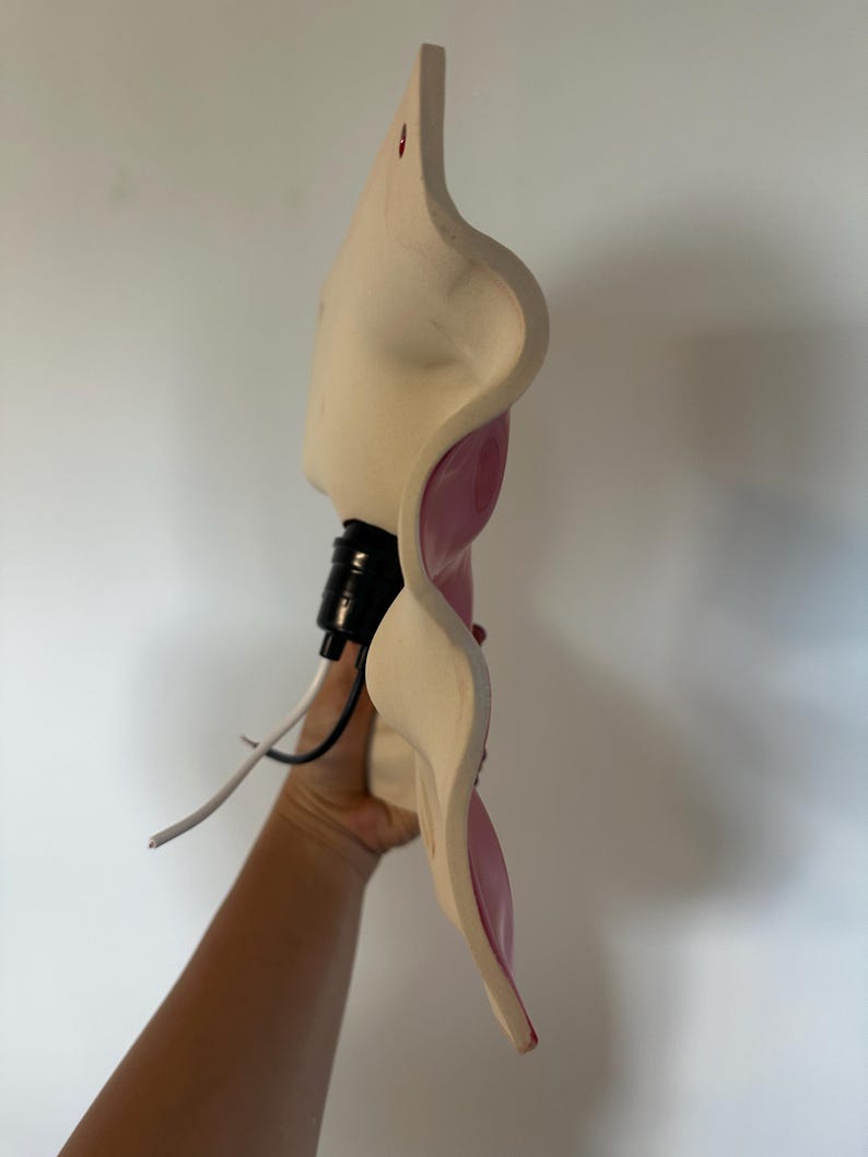 May include: A wall-mounted light fixture with a wavy, cream-colored ceramic base and a glossy pink interior surface. The fixture features a black socket extending from the center, with one white and one black wire protruding from the base.