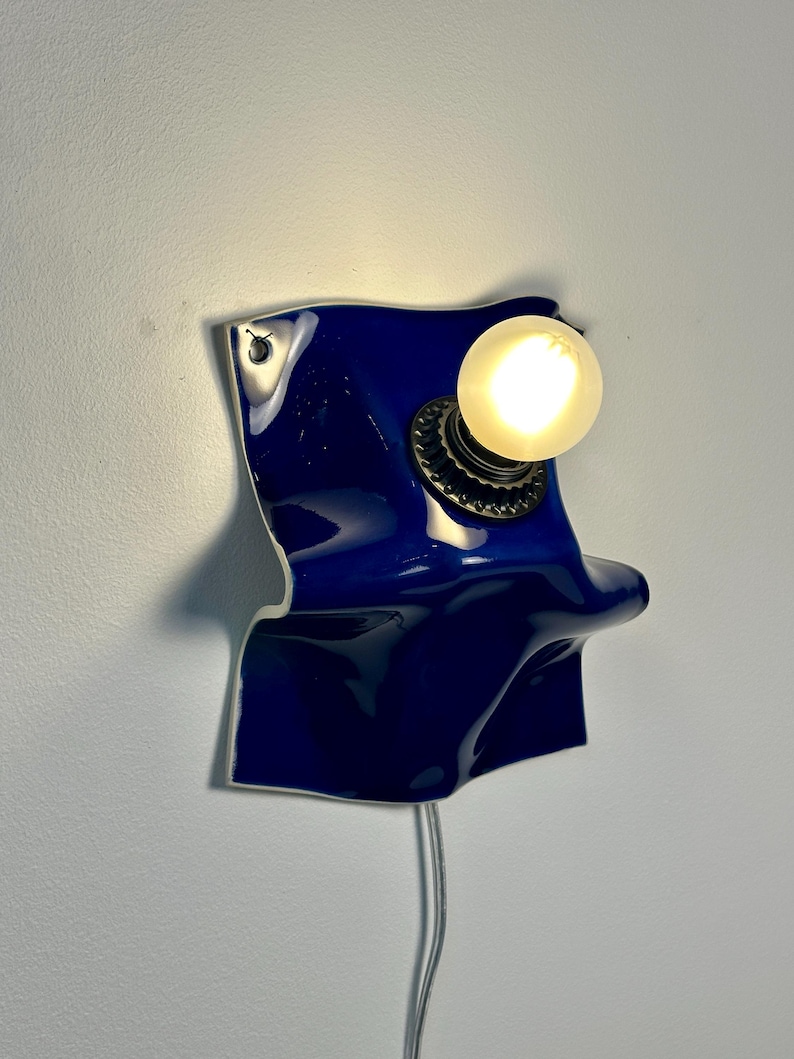 May include: A wall-mounted lamp featuring a glossy, deep blue, wavy ceramic base with an irregular, folded shape. A single, round, frosted light bulb is attached to the center of the blue surface, emitting a warm glow. A thin power cord hangs below.
