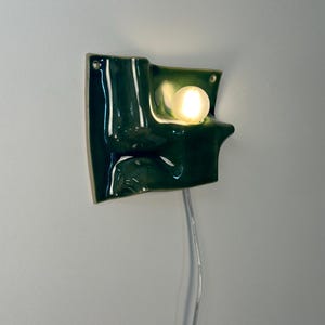 May include: A wall-mounted ceramic light fixture with a glossy, dark green, undulating surface. The piece features a single, round, frosted glass bulb emitting a warm glow, integrated into the curved form. A thin, clear power cord hangs downward.
