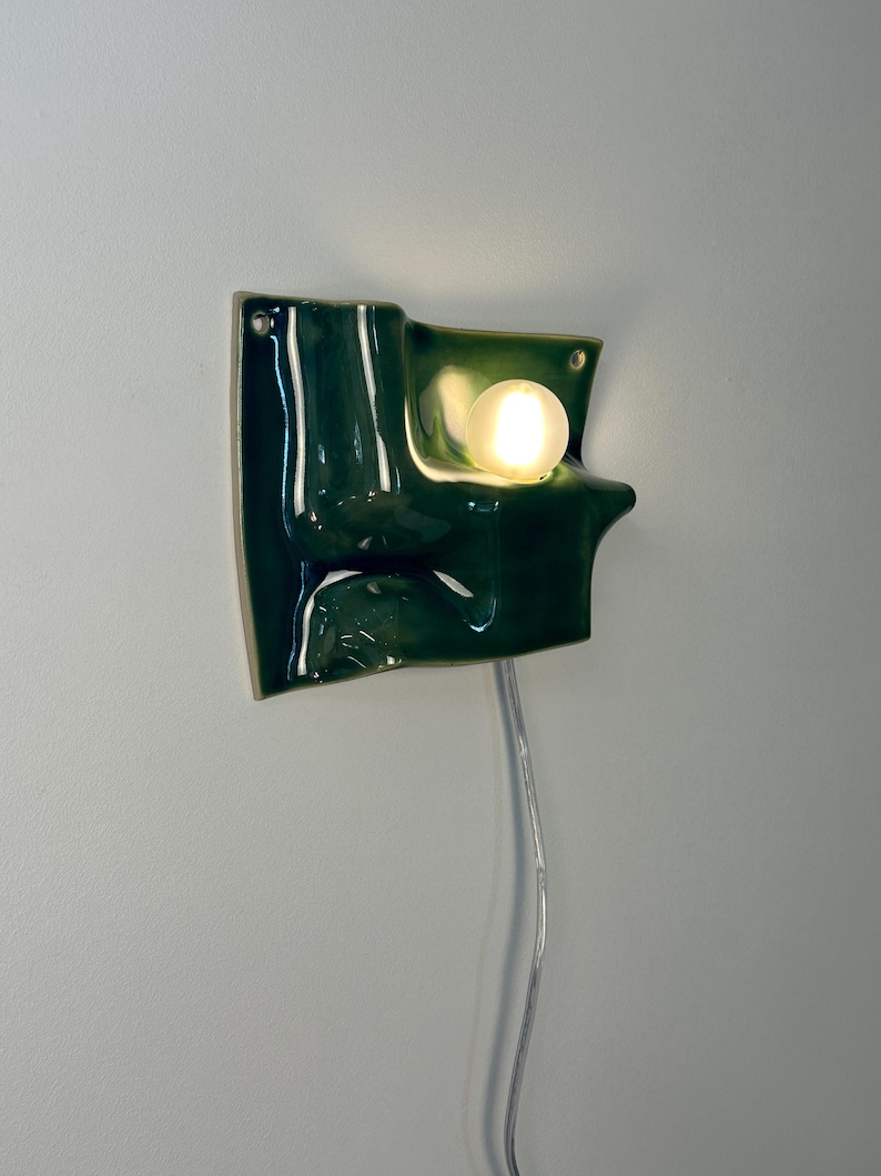 May include: A wall-mounted ceramic light fixture with a glossy, dark green, undulating surface. The piece features a single, round, frosted glass bulb emitting a warm glow, integrated into the curved form. A thin, clear power cord hangs downward.