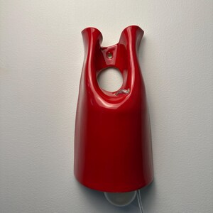 May include: A glossy red ceramic wall sconce featuring a smooth, curved, bell-like silhouette with a central circular cutout near the top. The fixture includes a mounting hole at the upper center and a white light bulb extending from the bottom edge.