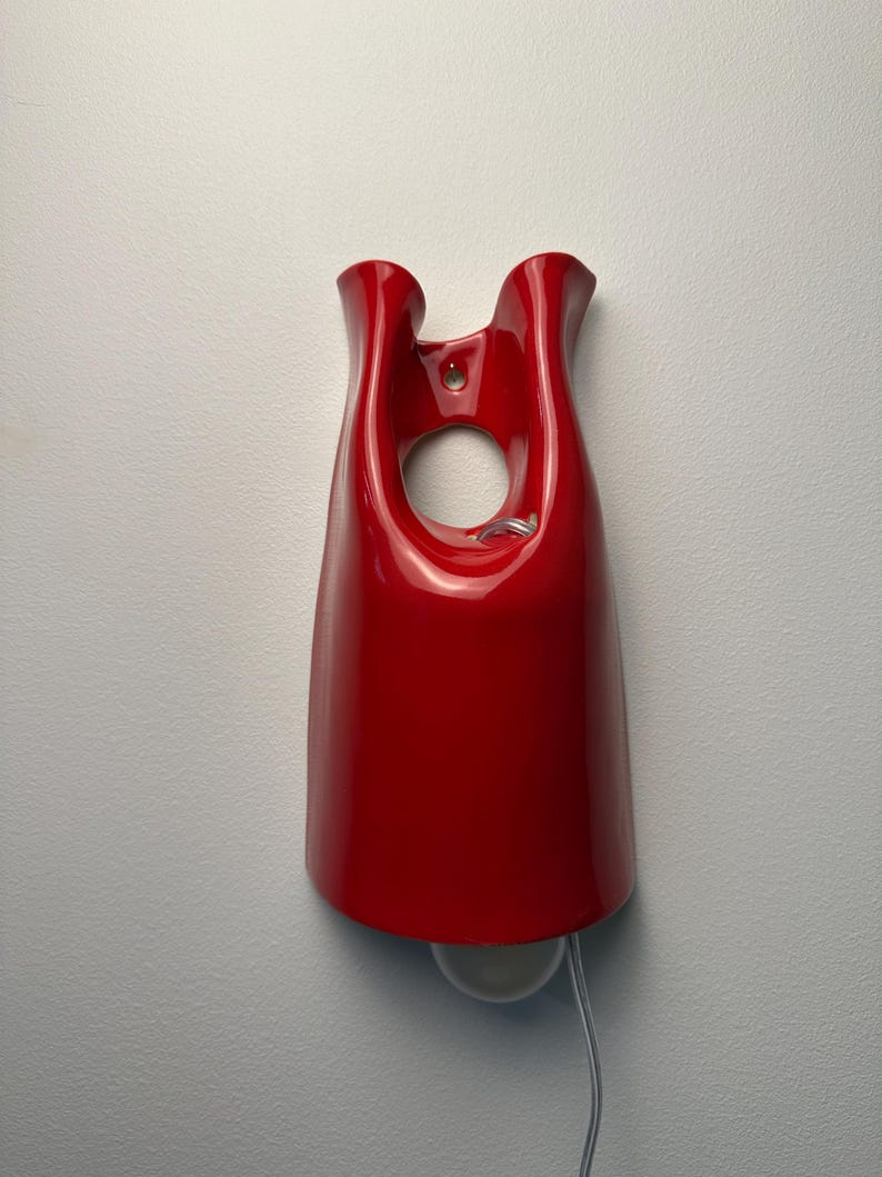May include: A glossy red ceramic wall sconce featuring a smooth, curved, bell-like silhouette with a central circular cutout near the top. The fixture includes a mounting hole at the upper center and a white light bulb extending from the bottom edge.
