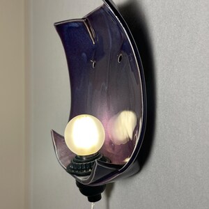 May include: A wall-mounted ceramic sconce featuring a curved, crescent-shaped backplate with a glossy, deep purple glaze. A single round, frosted light bulb is secured in a black socket at the base, emitting a warm glow against the purple surface.