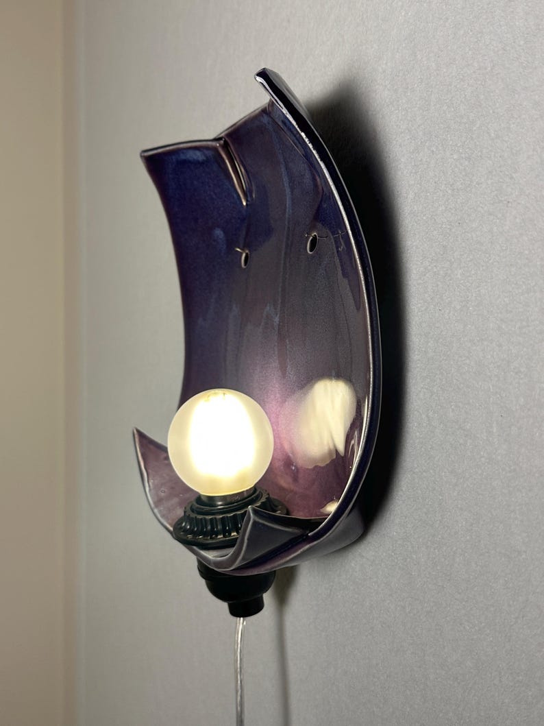 May include: A wall-mounted ceramic sconce featuring a curved, crescent-shaped backplate with a glossy, deep purple glaze. A single round, frosted light bulb is secured in a black socket at the base, emitting a warm glow against the purple surface.