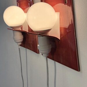 Puede incluir: Dos apliques de pared con placas traseras rectangulares de cer&aacute;mica roja oscura brillante. Cada l&aacute;mpara tiene un casquillo blanco con una bombilla redonda de luz blanca y un cable de alimentaci&oacute;n fino y transparente hacia abajo.