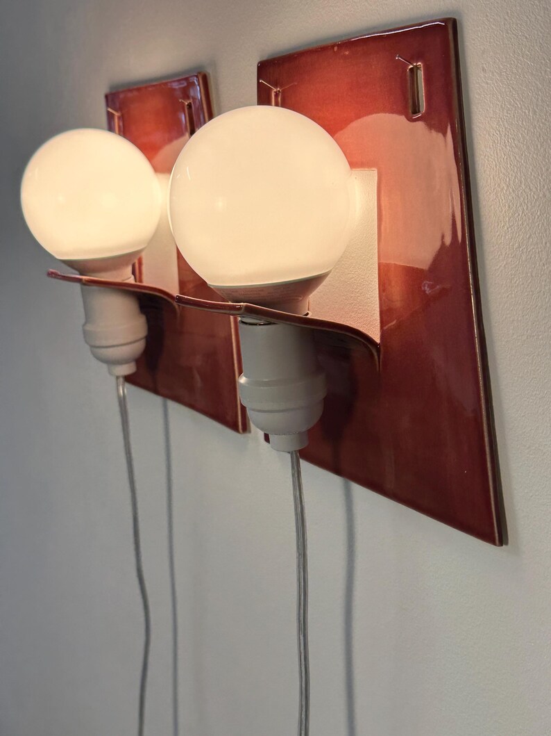 Puede incluir: Dos apliques de pared con placas traseras rectangulares de cer&aacute;mica roja oscura brillante. Cada l&aacute;mpara tiene un casquillo blanco con una bombilla redonda de luz blanca y un cable de alimentaci&oacute;n fino y transparente hacia abajo.