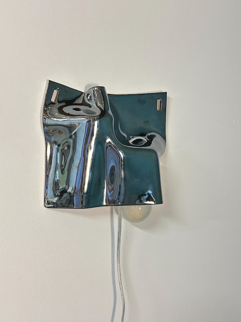 May include: A wall-mounted lamp featuring a reflective, chrome-finished metal shade with an irregular, fluid, and undulating surface. The fixture includes a single exposed light bulb extending from the bottom and a thin, clear power cord.