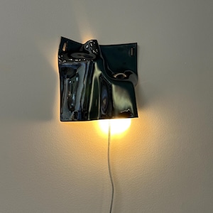 May include: A wall-mounted lamp featuring a glossy, dark, undulating ceramic shade with a sculptural, folded form. The fixture emits a warm, downward-facing light from a circular bulb, with a thin power cord extending from the bottom edge.