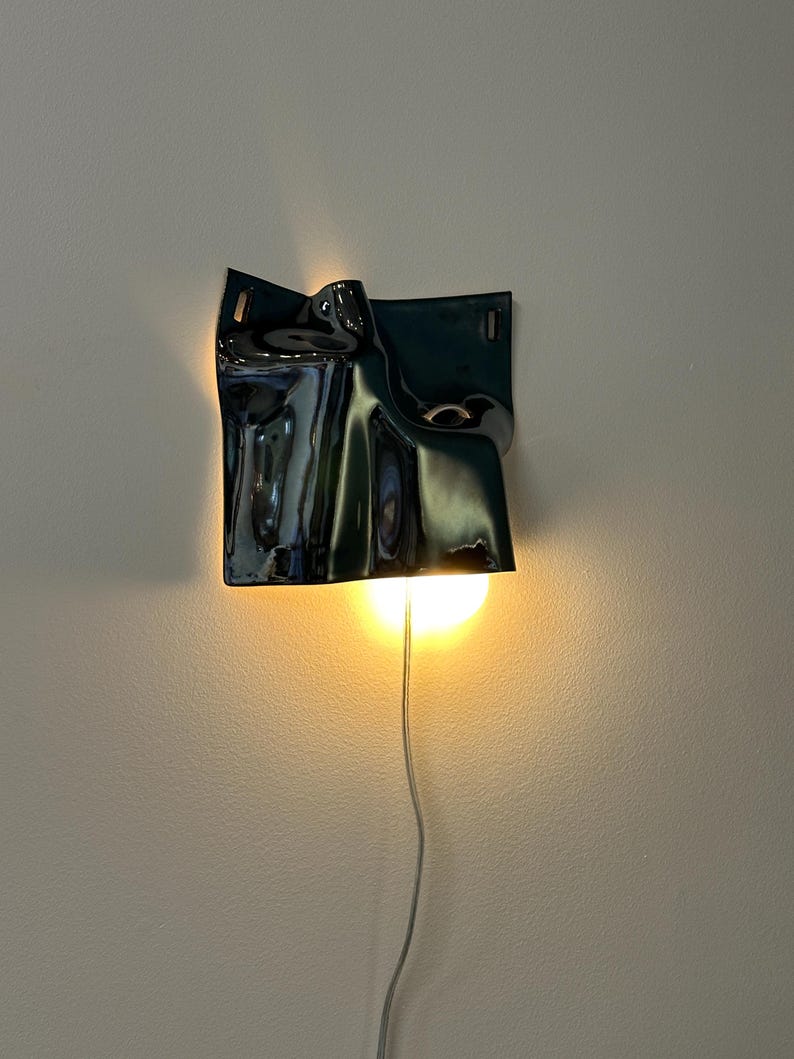 May include: A wall-mounted lamp featuring a glossy, dark, undulating ceramic shade with a sculptural, folded form. The fixture emits a warm, downward-facing light from a circular bulb, with a thin power cord extending from the bottom edge.