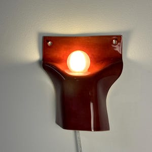 Pode incluir: Um candeeiro de parede em cer&acirc;mica com um acabamento vidrado vermelho profundo e brilhante. A pe&ccedil;a apresenta uma forma org&acirc;nica curva com dois orif&iacute;cios de montagem circulares nos cantos superiores. Uma l&acirc;mpada redonda de vidro fosco emite luz.